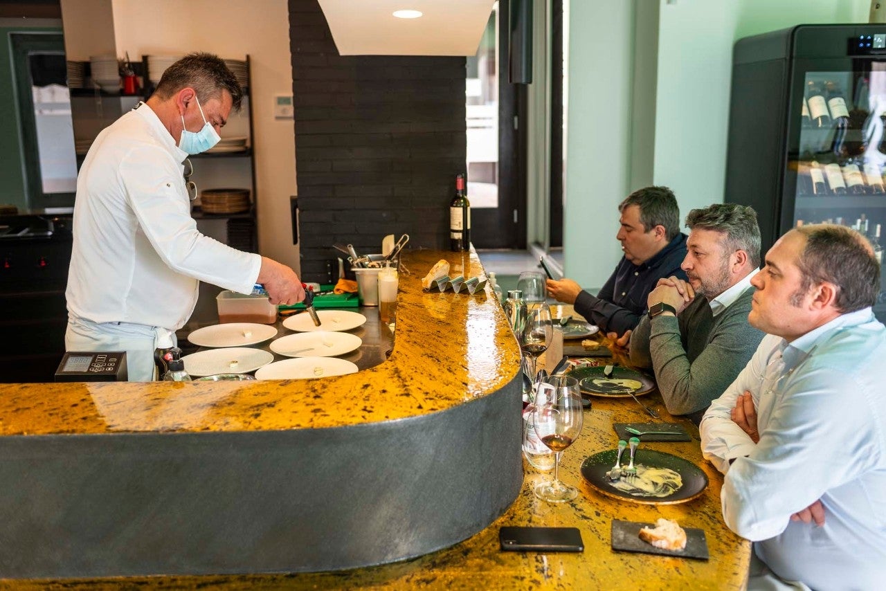 El cocinero elabora varios platos en la barra japonesa frente a tres comensales.