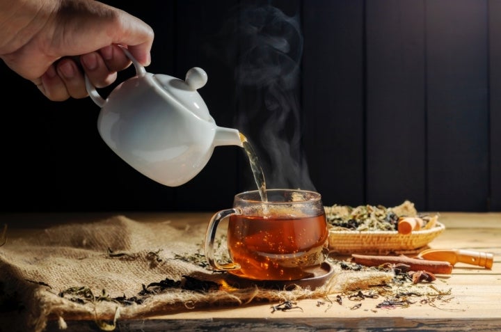 Pour hot tea into cups, herbal tea glasses are arranged on a rustic wooden table.
