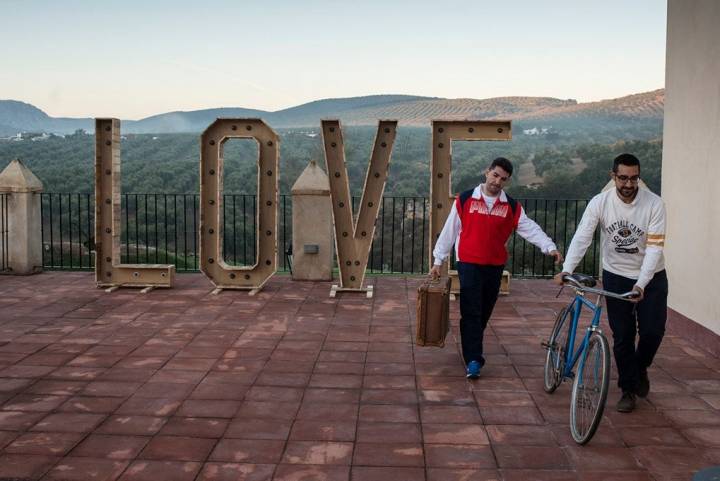 Dos hombres: uno llevando una bicicleta y el otro una maleta en una terraza donde hay unas letras que forman la palabra "love"