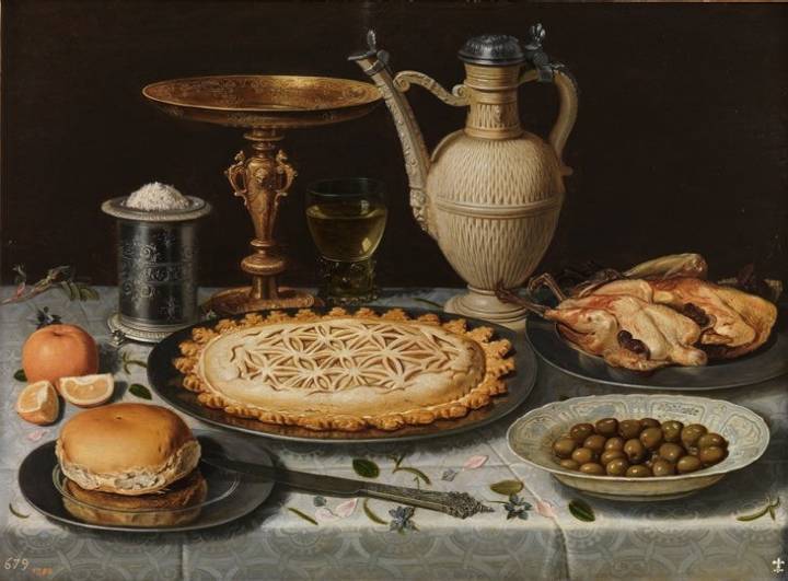 Mesa con mantel, salero, taza dorada, pastel, jarra, plato de porcelana con aceitunas y aves asadas.