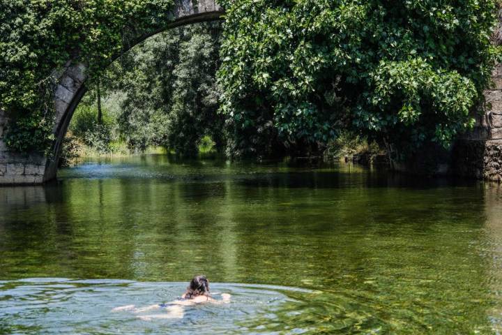 Mujer nadando en las aguas de la piscina natural de Carreciá, con el puente y la vegetación frondosa al fondo.